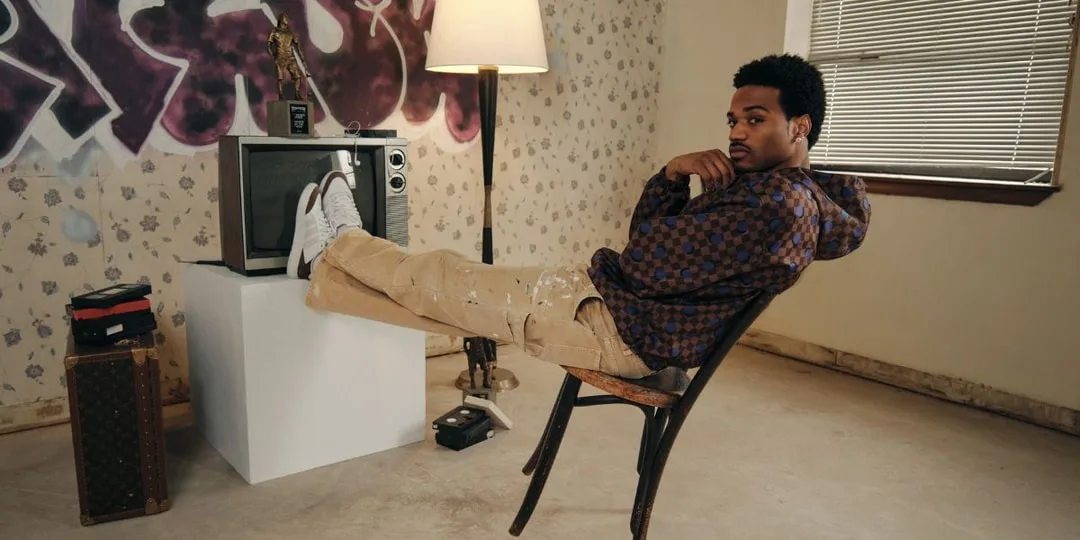 Homme assis sur une chaise, pieds sur une TV, graffiti en arrière-plan.