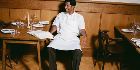 Un serveur en tablier blanc sourit, assis dans un restaurant en bois.
