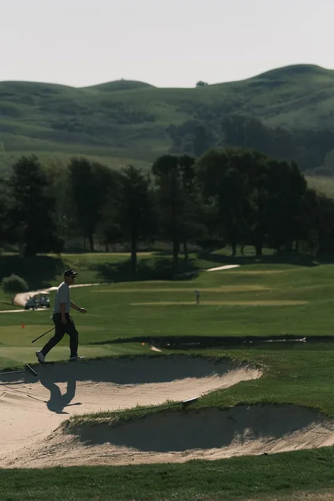 Golfeur marchant près d'un bunker sur un parcours de golf vallonné.