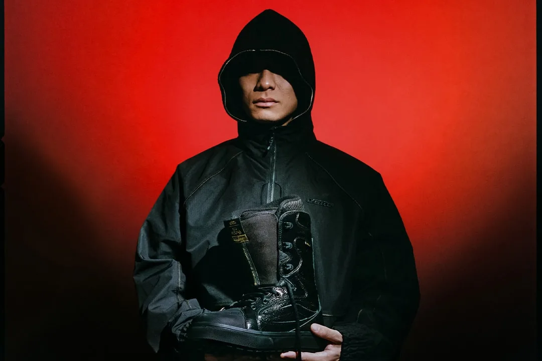 Personne en noir avec un sweat à capuche tenant une botte noire, fond rouge.