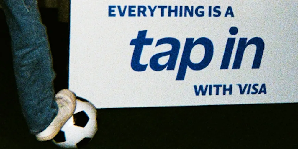 Pied sur un ballon de football à côté d'une affiche 'EVERYTHING IS A tap in WITH VISA'.