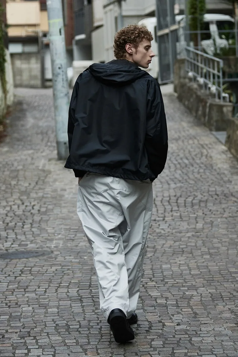 Homme portant une veste noire et pantalon blanc, marchant dans une ruelle pavée.