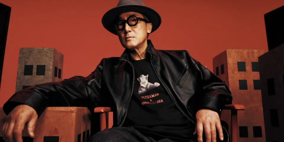 Un homme en chapeau noir et lunettes est assis devant un décor rouge.