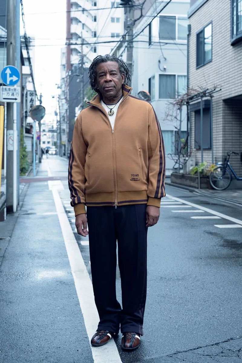 Un homme debout dans une rue de ville, vêtu d'une veste marron et pantalon noir.