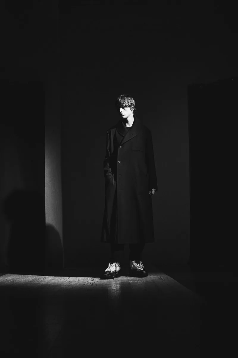 Homme en manteau long dans un éclairage dramatique en noir et blanc.
