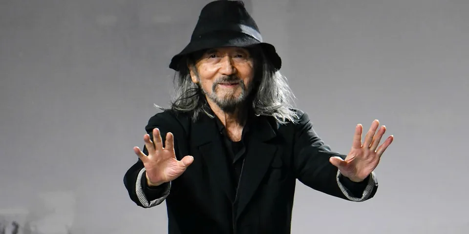 Homme aux cheveux longs, vêtu de noir et coiffé d’un chapeau, les mains levées.